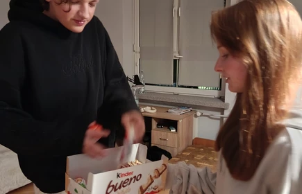 Na zdjęciu widać dwie osoby stojące w pokoju. Osoba po lewej, ubrana w czarną bluzę, trzyma pudełko z napisem „Kinder”. Osoba po prawej, w jasnej bluzie, sięga po coś z pudełka.
