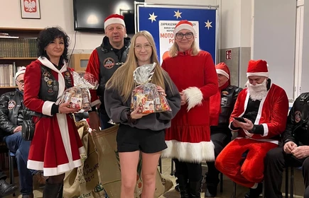 Na zdjęciu widać grupę os&oacute;b podczas świątecznego spotkania. Kilka z nich jest ubranych w stroje Mikołaja lub w czerwono-białe stylizacje. Wszyscy wyglądają na uśmiechniętych i trzymają paczki z upominkami, co sugeruje akcję charytatywną lub mikołajkową. Scena odbywa się w pomieszczeniu z dekoracjami świątecznymi.