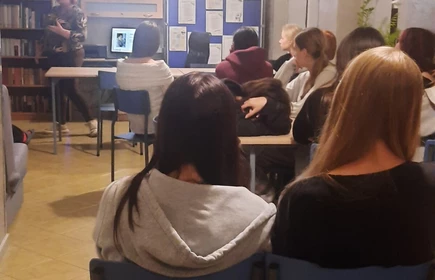  siedzący uczniowie przy stołach, zwróconych w stronę prowadzącej, która stoi z przodu pomieszczenia.