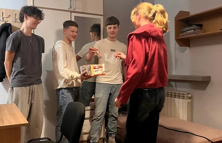 Na zdjęciu widać cztery młode osoby stojące , które rozmawiają i spędzają razem czas w swobodnej atmosferze. Jedna osoba ma na sobie czerwoną bluzę z kapturem i stoi bliżej prawej strony, zwrócona w stronę pozostałych,jedna osoba w jasnym swetrze trzyma coś w rękach najprawdopodobniej słodycze