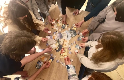 Na zdjęciu widać grupę młodych os&oacute;b stojących wok&oacute;ł drewnianego stołu. Wszyscy pochylają się nad stołem i trzymają w rękach kolorowe karty oraz okrągłe przypinki lub żetony