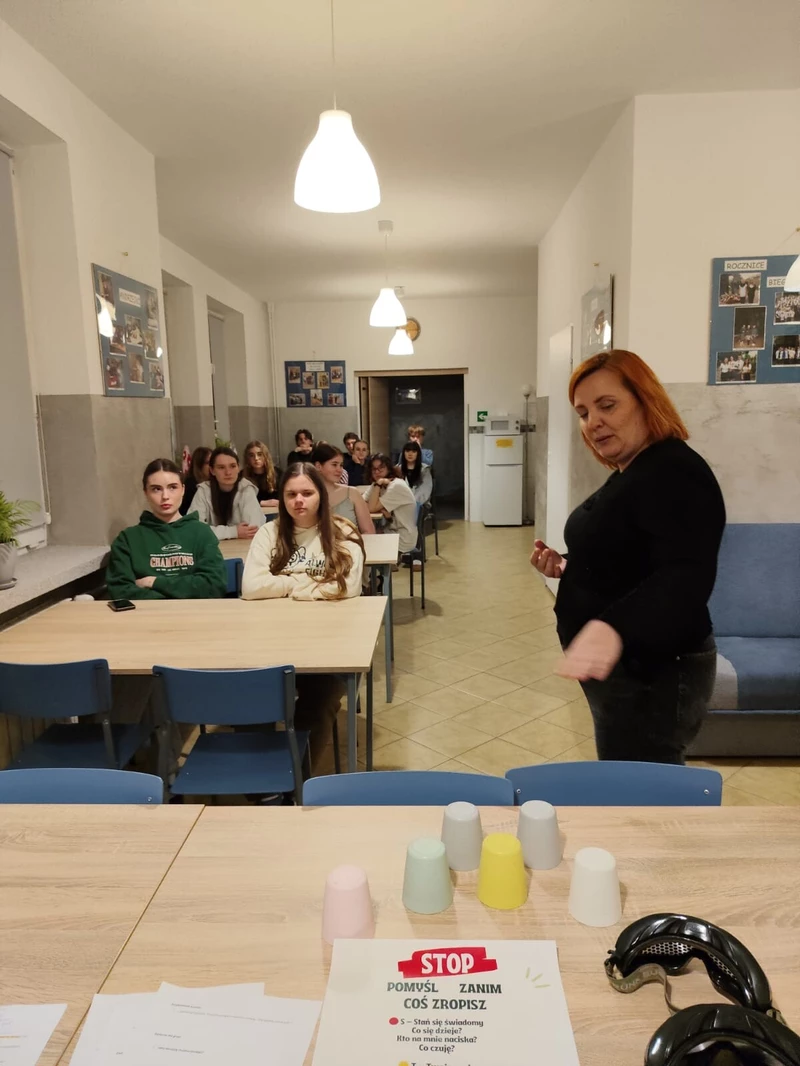 Na zdjęciu widać salę przypominającą szkolną świetlicę lub klasę. Przy kilku ustawionych w rzędach stołach siedzi grupa nastolatek, które patrzą w stronę przodu pomieszczenia. Ich wyrazy twarzy są raczej poważne i skupione.

Na pierwszym planie, po prawej stronie, stoi dorosła kobieta (prawdopodobnie nauczycielka lub prowadząca zajęcia). Jest odwrócona częściowo bokiem i wykonuje gest ręką, jakby coś tłumaczyła albo instruowała uczestników.