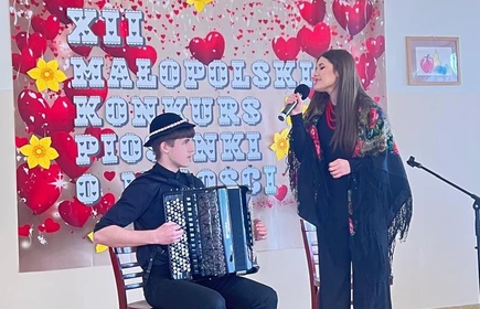 na zdjęciu widoczne są dwie osoby chłopiec siedzący na krzesle  na głowie ma kapelusz a w rekach trzyma akordeon  jest skierowany do stojącej dziewczyny kt&oacute;ra w prawej ręce trzyma mikrofon obydwoje stoją przed plakatem zawisnietym na białej scianie na kt&oacute;rym widnieje napis XII Małopolski Konkurs Piosenki o Miłości 