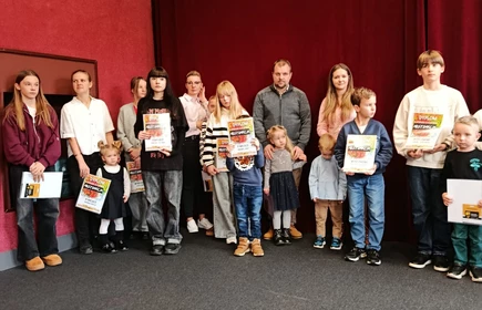 W tle znajduje się grupa uśmiechniętych osób, najpewniej świadków uroczystości  wręczenia dyp— mogą to być nauczyciele lub inni uczestnicy wydarzenia. Całość odbywa się w miejscu z czerwonymi zasłonami, co nadaje atmosferę szkolnej lub lokalnej gali. Wszyscy wyglądają na zadowolonych i dumnych z tego momentu.