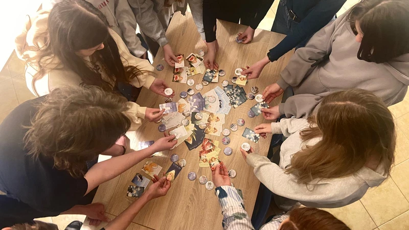 Na zdjęciu widać grupę młodych osób stojących wokół drewnianego stołu. Wszyscy pochylają się nad stołem i trzymają w rękach kolorowe karty oraz okrągłe przypinki lub żetony