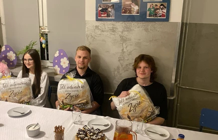 Na zdjęciu widać cztery osoby siedzące przy stole w sali, kt&oacute;ra wygląda jak szkolna lub świetlicowa. Każda z nich trzyma zapakowany prezent z napisem &bdquo;Idź odważnie przez swoje życie&rdquo;. Na stole stoją talerzyki, kubki, ciasto, sok i przekąski. W tle są dekoracje z wielkanocnymi jajkami oraz tablica ze zdjęciami. Atmosfera wygląda na uroczystą i miłą