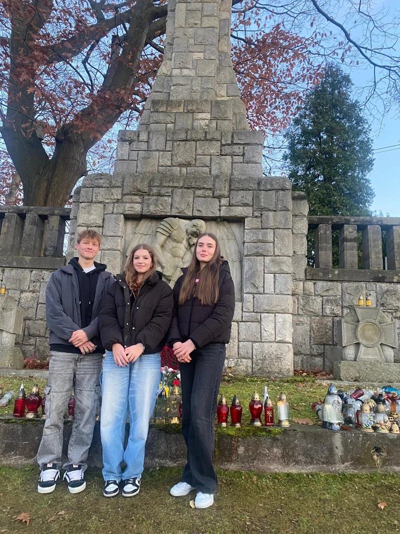 Na zdjęciu stoją trzy osoby przed kamiennym pomnikiem. Mają na sobie ciepłe ubrania, a przed nimi ustawione są znicze i kwiaty. Scena wygląda na spokojną i upamiętniającą.