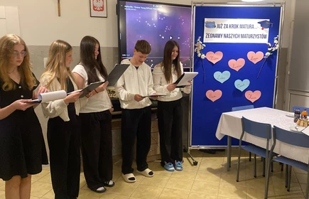 Na zdjęciu sześć os&oacute;b stoi obok siebie i czyta z kartek, jak podczas kr&oacute;tkiego występu. W tle widać ekran z włączonym teledyskiem oraz tablicę z napisem &bdquo;Już za krok matura&hellip; Żegnamy naszych maturzyst&oacute;w&rdquo; i papierowymi sercami z imionami. Obok stoi st&oacute;ł z ciastem i napojami