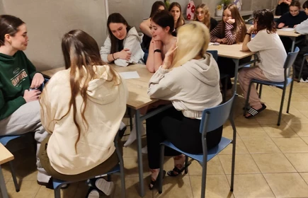 Na zdjęciu widać grupę młodych os&oacute;b siedzących przy stołach w sali przypominającej klasę lub świetlicę. Siedzą w kilkuosobowych grupach, rozmawiają i pracują nad czymś na kartkach leżących na stole.

Atmosfera wygląda na swobodną i koleżeńską &mdash; część os&oacute;b się uśmiecha, inni słuchają lub są skupieni. W tle widać ściany z plakatami i zdjęciami oraz okna, co sugeruje szkolne lub warsztatowe zajęcia grupowe.
