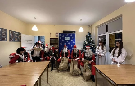 Siedmiu siedzących  Mikołajów z prezentami obok wychowawczyni i stojące uczennice przebrane za Anioły