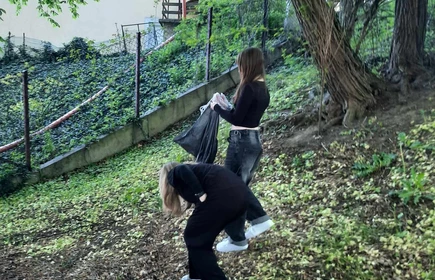 pochylona uczennica podnosząca  śmieci i stojąca obok   uczennica trzymająca worek do którego zbierane są smieci 