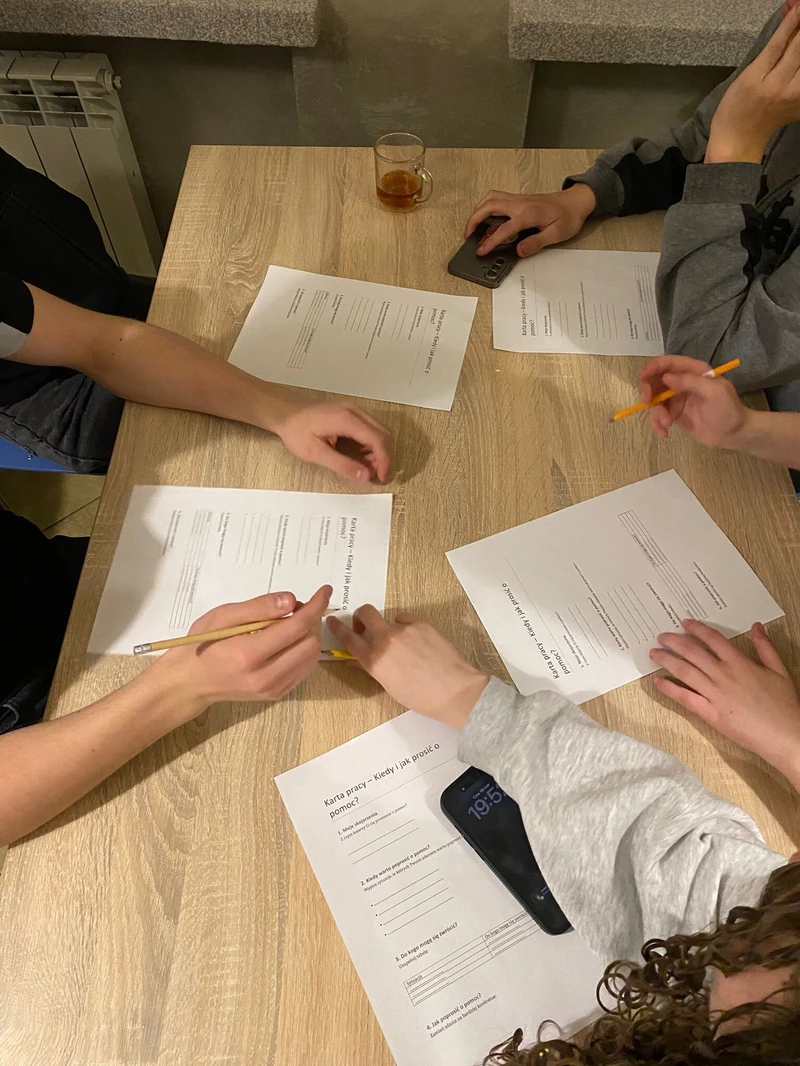 na zdjęciu widac blat drewniany na ktorym oparte są ręce uczniów a przy nich karty pracy