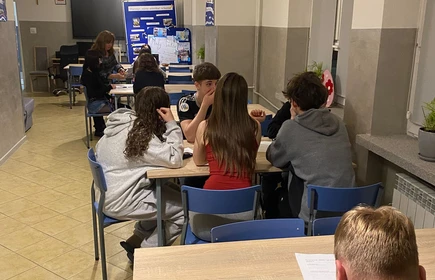 zdjęcie przedstawia siedzących uczni&oacute;w przy stolikach podczas warsztat&oacute;w