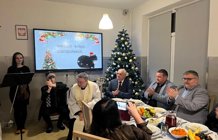 na zdjęciu widać monitor z napisem wiecz&oacute;r Kolęd przed nim st&oacute;ł wigilijny przy kt&oacute;rym siedzą osoby  dorosłe  i stojąca uczennica czytającą tekst