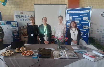 zdjęcie przedstawia st&oacute;ł z rekwizytami promującymi plac&oacute;wkę ,przed stołem stoją uczniowie wraz z nauczycielem