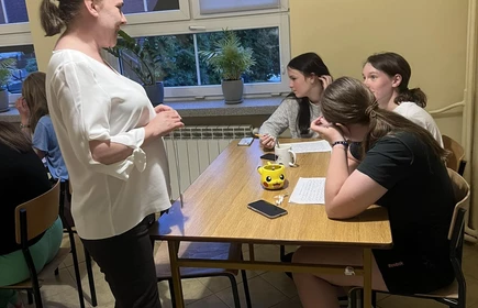 prowadząca stojąca nad stołem przy którym siedzą uczniowie