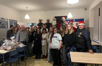 Na zdjęciu widać grupę os&oacute;b podczas świątecznego spotkania. Kilka z nich jest ubranych w stroje Mikołaja lub w czerwono-białe stylizacje. Wszyscy wyglądają na uśmiechniętych i trzymają paczki z upominkami, co sugeruje akcję charytatywną lub mikołajkową. Scena odbywa się w pomieszczeniu z dekoracjami świątecznymi.