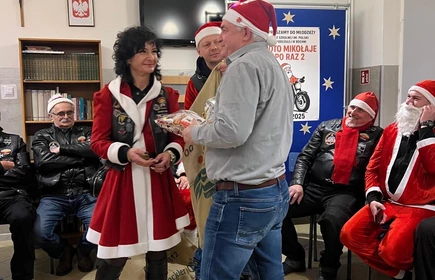 Na zdjęciu widać grupę os&oacute;b podczas świątecznego spotkania. Kilka z nich jest ubranych w stroje Mikołaja lub w czerwono-białe stylizacje. Wszyscy wyglądają na uśmiechniętych i trzymają paczki z upominkami, co sugeruje akcję charytatywną lub mikołajkową. Scena odbywa się w pomieszczeniu z dekoracjami świątecznymi.