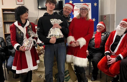 Na zdjęciu widać grupę os&oacute;b podczas świątecznego spotkania. Kilka z nich jest ubranych w stroje Mikołaja lub w czerwono-białe stylizacje. Wszyscy wyglądają na uśmiechniętych i trzymają paczki z upominkami, co sugeruje akcję charytatywną lub mikołajkową. Scena odbywa się w pomieszczeniu z dekoracjami świątecznymi.