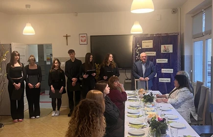 Na zdjęciu widać w oddali stojacych uczni&oacute;w przedstawiajacych uroczystosc wielkanocną ,następnie przy stole  na ktorym dominują kolory zielone i ż&oacute;łte, kt&oacute;re mogą sugerować trawę oraz drobne kwiaty lub rośliny,siędza uczniowie  przygotowujący się do spożycia posiłku
