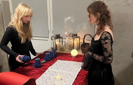 Dwie  dziewczyny stoja na przeciw siebie przy stole na którym są gadżety andrzejkowe