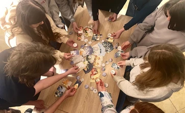 Na zdjęciu widać grupę młodych osób stojących wokół drewnianego stołu. Wszyscy pochylają się nad stołem i trzymają w rękach kolorowe karty oraz okrągłe przypinki lub żetony
