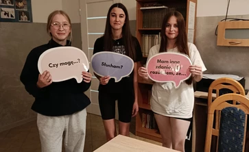 Na zdjęciu widać trzy młode osoby stojące w pomieszczeniu, prawdopodobnie w sali szkolnej lub świetlicy. Każda z nich trzyma w rękach tabliczkę w kształcie dymku z napisem:
pierwsza osoba z lewej ma napis „Cześć!”,
środkowa osoba — „Dziękuję!”,
osoba po prawej — „Proszę!”.