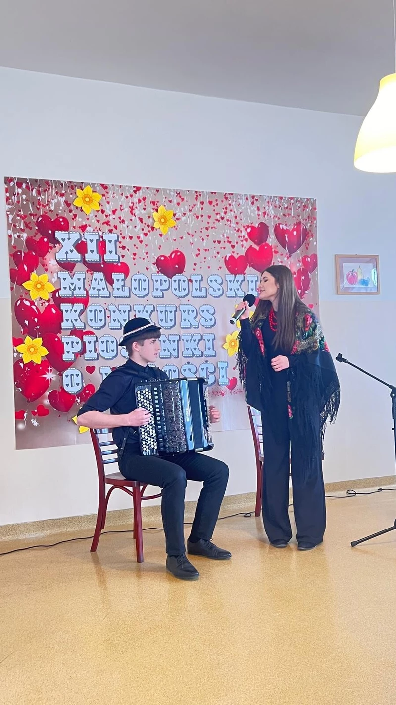 na zdjęciu widoczne są dwie osoby chłopiec siedzący na krzesle  na głowie ma kapelusz a w rekach trzyma akordeon  jest skierowany do stojącej dziewczyny która w prawej ręce trzyma mikrofon obydwoje stoją przed plakatem zawisnietym na białej scianie na którym widnieje napis XII Małopolski Konkurs Piosenki o Miłości 