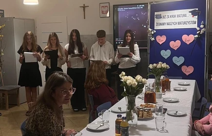 Na zdjęciu widać długi st&oacute;ł nakryty do poczęstunku z ciastem, napojami i kwiatami. Przy stole siedzą osoby, a z tyłu kilka os&oacute;b stoi i czyta z kartek podczas występu. Obok jest tablica z napisem o pożegnaniu maturzyst&oacute;w.