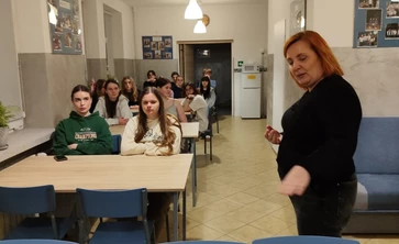 Na zdjęciu widać salę przypominającą szkolną świetlicę lub klasę. Przy kilku ustawionych w rzędach stołach siedzi grupa nastolatek, które patrzą w stronę przodu pomieszczenia. Ich wyrazy twarzy są raczej poważne i skupione.

Na pierwszym planie, po prawej stronie, stoi dorosła kobieta (prawdopodobnie nauczycielka lub prowadząca zajęcia). Jest odwrócona częściowo bokiem i wykonuje gest ręką, jakby coś tłumaczyła albo instruowała uczestników.