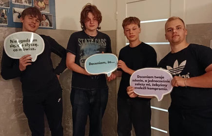 Na zdjęciu widać cztery młode osoby stojące w pomieszczeniu,  Każda z nich trzyma w rękach tabliczkę w kształcie dymku z napisem