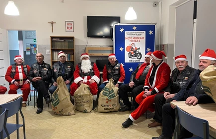 Na zdjęciu widać grupę os&oacute;b podczas świątecznego spotkania. Kilka z nich jest ubranych w stroje Mikołaja lub w czerwono-białe stylizacje. Wszyscy wyglądają na uśmiechniętych i trzymają paczki z upominkami, co sugeruje akcję charytatywną lub mikołajkową. Scena odbywa się w pomieszczeniu z dekoracjami świątecznymi.