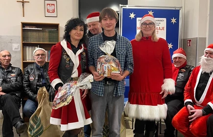 Na zdjęciu widać grupę os&oacute;b podczas świątecznego spotkania. Kilka z nich jest ubranych w stroje Mikołaja lub w czerwono-białe stylizacje. Wszyscy wyglądają na uśmiechniętych i trzymają paczki z upominkami, co sugeruje akcję charytatywną lub mikołajkową. Scena odbywa się w pomieszczeniu z dekoracjami świątecznymi.