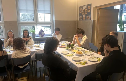 Na zdjęciu widać grupę uczni&oacute;w siedzących przy stole wielkanocnym podczas uroczystego spotkania z okazji Wielkanoc Na stole stoją talerze z jedzeniem, przekąski oraz świąteczne dekoracje, co podkreśla uroczysty charakter spotkania. Wszyscy są elegancko ubrani i zwracają uwagę w stronę os&oacute;b stojących z przodu sali.