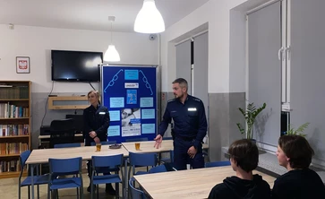 Przy kilku ustawionych obok siebie stołach siedzi jedna osoba (widoczna od tyłu), a dwie inne stoją przy stołach. Te dwie osoby mają na sobie ciemne, jednakowe stroje służbowe, co sugeruje, że mogą prowadzić zajęcia, spotkanie lub prezentację.

W tle stoi tablica ekspozycyjna z kilkoma plakatami i wydrukami, które wyglądają na materiały edukacyjne.