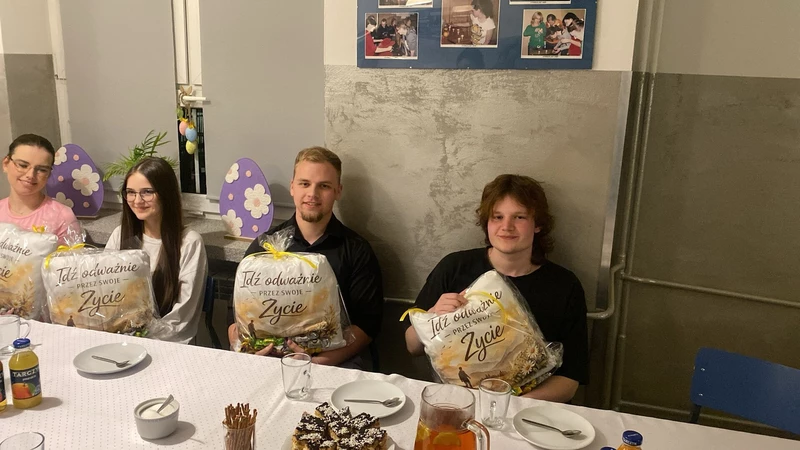 Na zdjęciu widać cztery osoby siedzące przy stole w sali, która wygląda jak szkolna lub świetlicowa. Każda z nich trzyma zapakowany prezent z napisem „Idź odważnie przez swoje życie”. Na stole stoją talerzyki, kubki, ciasto, sok i przekąski. W tle są dekoracje z wielkanocnymi jajkami oraz tablica ze zdjęciami. Atmosfera wygląda na uroczystą i miłą