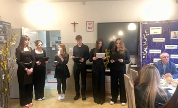 Po lewej stronie stoją  dziewczyny ubrane na czarno i i chłopiec trzymają kartki  i czytają tekst z kartek i prezentują krótką część artystyczną

Za nimi znajduje się dekoracyjna plansza w fioletowym kolorze. Widać na niej napisy: „Wielkanoc 2026”, „Nadzieja”, „Rodzina”, „Przebaczenie”, „Radość”. Ozdobiona jest też wiosennymi elementami.Po prawej stronie  widac dwie dorosłe osoby
