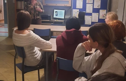  Grupa osób uczestniczących w spotkaniu lub zajęciach w sali edukacyjnej. Kilka osób siedzi przy stołach, zwróconych w stronę prowadzącej, która stoi z przodu pomieszczenia. Kobieta prowadząca prezentację gestykuluje ręką i mówi do uczestników.