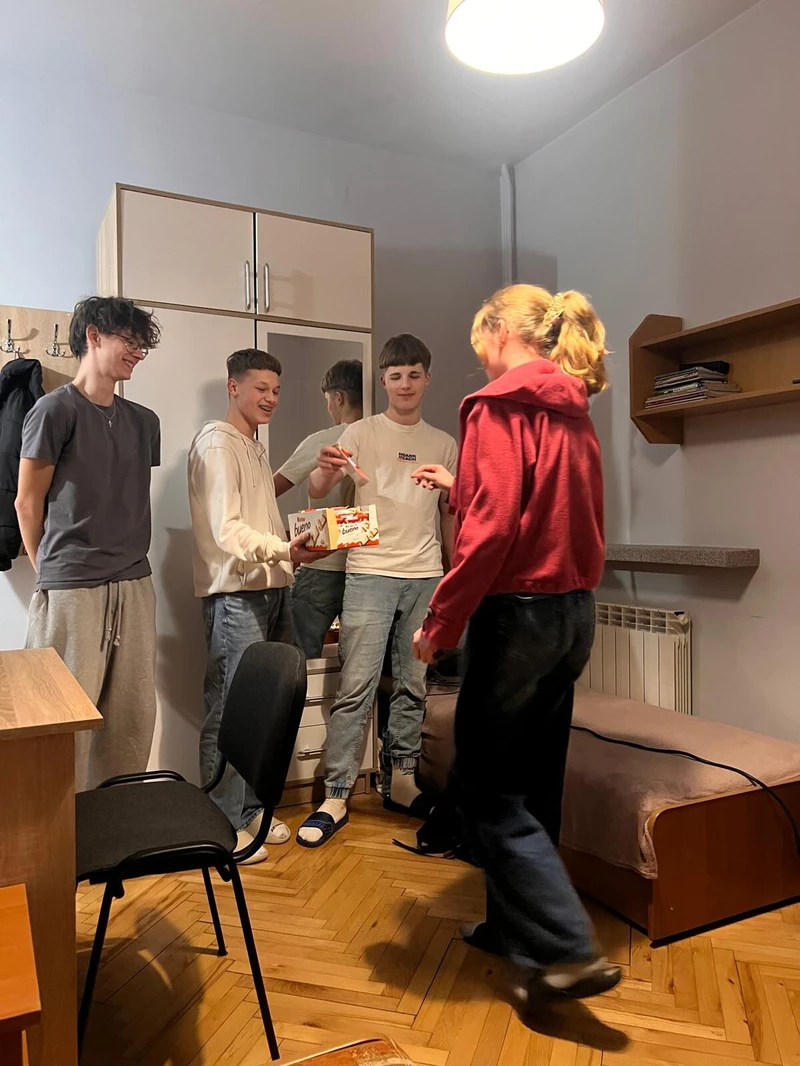 Na zdjęciu widać cztery młode osoby stojące , które rozmawiają i spędzają razem czas w swobodnej atmosferze. Jedna osoba ma na sobie czerwoną bluzę z kapturem i stoi bliżej prawej strony, zwrócona w stronę pozostałych,jedna osoba w jasnym swetrze trzyma coś w rękach najprawdopodobniej słodycze