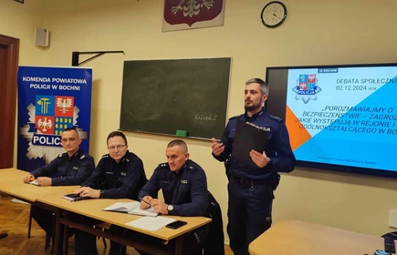 stojący policjant i siedzący obok niego przy stole policjanci wygłaszający prelekcje o bezpieczeństwie
