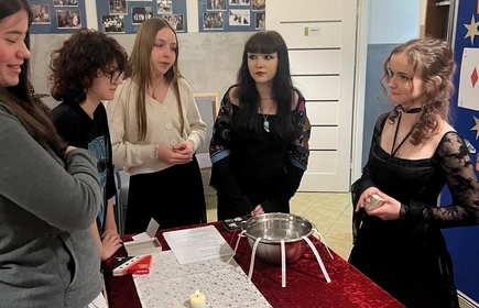 Cztery uczennice stoją obok siebie, są uśmiechnięte Stoją obok siebie, uśmiechnięte 
Przed nimi stoi stół nakryty czerwonym materiałem i jasnym bieżnikiem. Na stole znajdują się przedmioty używane podczas zabaw andrzejkowych — m.in. miska z  z której wystają paski z napisami