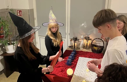 Przy stole  z gadżetami  wróżb andrzejkowych  stoja osoby -widoczne trzy dziewczyny jeden chłopiec. Dziewczyny maja na głowie kapelusze