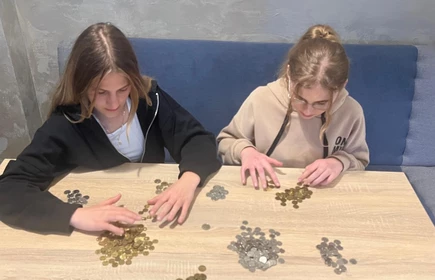 na zdjęciu widac dwie uczennice siedzace przy stole na kt&oacute;rym są ułożone stosy  z monetami