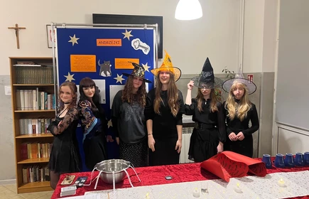 ChatGPT powiedział:

Na zdjęciu widać grupę sześciu młodych osób przebranych w stroje inspirowane andrzejkowymi wróżbami — wszystkie są ubrane na czarno, a część z nich ma na głowach ozdobne kapelusze czarownic w różnych kolorach i wzorach. Stoją obok siebie, uśmiechnięte lub pozujące, na tle dużej niebieskiej tablicy dekoracyjnej z napisem „ANDRZEJKI”, gwiazdkami oraz ilustracjami związanymi z tradycyjnymi wróżbami.