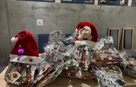 Na zdjęciu widać grupę os&oacute;b podczas świątecznego spotkania. Kilka z nich jest ubranych w stroje Mikołaja lub w czerwono-białe stylizacje. Wszyscy wyglądają na uśmiechniętych i trzymają paczki z upominkami, co sugeruje akcję charytatywną lub mikołajkową. Scena odbywa się w pomieszczeniu z dekoracjami świątecznymi.