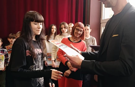 Na zdjęciu widać scenę wręczania nagrody lub dyplomu. Po lewej stronie stoi młoda osoba z ciemnymi, długimi włosami, ubrana w czarną bluzę. Odbiera dokument trzymany przez wysokiego mężczyznę w eleganckim, ciemnym ubraniu, stojącego po prawej stronie. W tle znajduje się grupa uśmiechniętych osób, najpewniej świadków uroczystości
