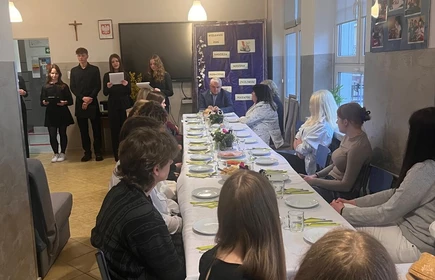 Na zdjęciu widać w oddali stojacych uczni&oacute;w przedstawiajacych uroczystosc wielkanocną ,następnie przy stole  na ktorym dominują kolory zielone i ż&oacute;łte, kt&oacute;re mogą sugerować trawę oraz drobne kwiaty lub rośliny,siędza uczniowie  przygotowujący się do spożycia posiłku
