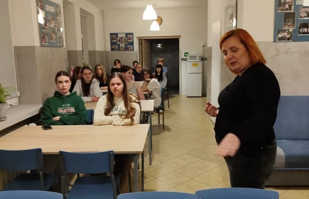 Na zdjęciu widać salę przypominającą szkolną świetlicę lub klasę. Przy kilku ustawionych w rzędach stołach siedzi grupa nastolatek, kt&oacute;re patrzą w stronę przodu pomieszczenia. Ich wyrazy twarzy są raczej poważne i skupione.

Na pierwszym planie, po prawej stronie, stoi dorosła kobieta (prawdopodobnie nauczycielka lub prowadząca zajęcia). Jest odwr&oacute;cona częściowo bokiem i wykonuje gest ręką, jakby coś tłumaczyła albo instruowała uczestnik&oacute;w.