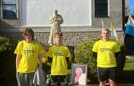 trzech uczniów w żółtych podkoszulkach  stojących przed pomnikiem Papieża