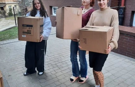 Po lewej stronie stoi dziewczyna z długimi, kręconymi włosami, ubrana w luźną bluzę i szerokie spodnie. W środku znajduje się druga osoba z rudymi włosami, ubrana w bordową koszulkę i dżinsy. Po prawej stronie stoi uśmiechnięta osoba w jasnym swetrze i sp&oacute;dnicy, mająca na nogach rajstopy w kropki i kapcie.Tło stanowi budynek bursy